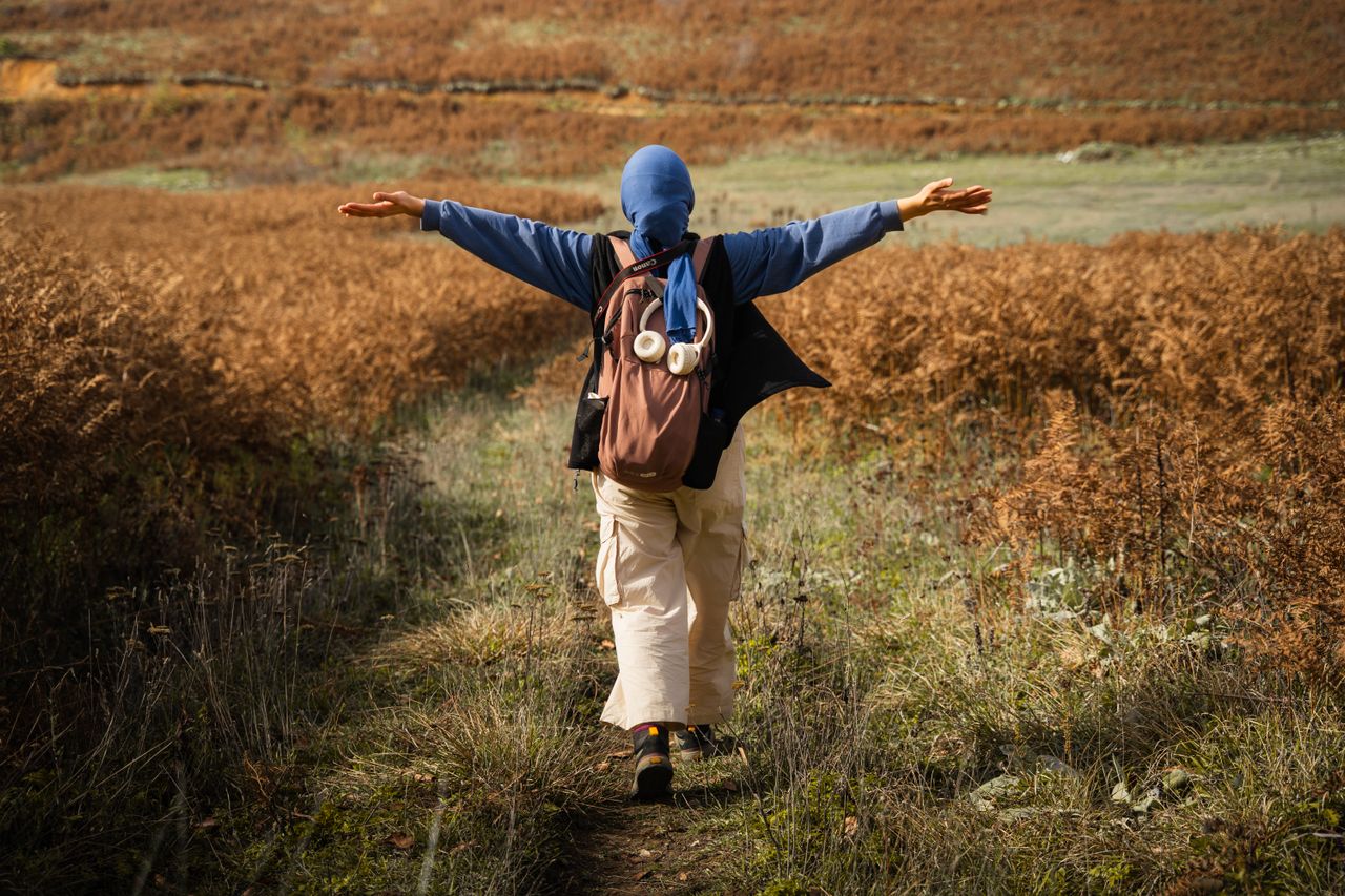 Woman with blue head scarf and backpack walking through field with arms out wide