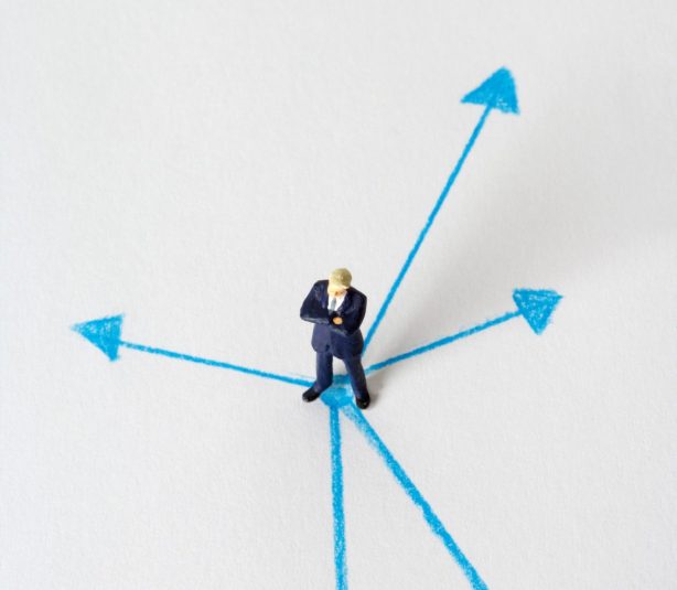 Miniature businessman stands at crossroads of blue arrows on white background, symbolizing choices.