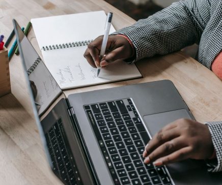 Women with open laptop and pad next to it writing with one hand and using laptop with other.