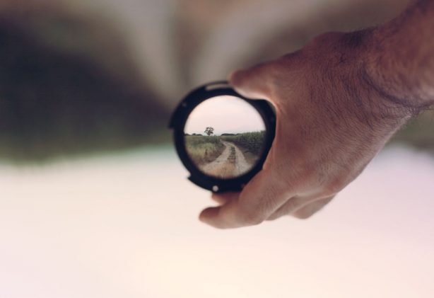 A hand holds a camera lens revealing a clear path through nature, symbolizing vision and focus.